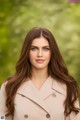 A woman with long brown hair wearing a beige coat.