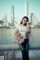 A woman in a fur coat leaning against a wall by the water.